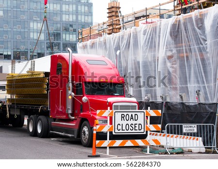 Red big rig American long haul semi truck with a long flat bed trailer unloads delivered for the construction of multi-storey building metal structures in a closed to traffic city street