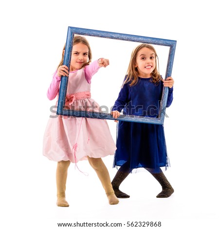 Identical twins girls are pointing with fingers holding picture frame. Children posing in studio, fooling around making different facial expressions and pointing at the camera with fingers.