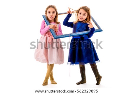 Identical twins girls are making happy expressions with picture frame. Children posing in studio, fooling around making different facial expressions.