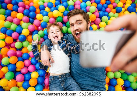 Cheerful joyful son and dad lying on colorful balls and taking selfie with smartphone