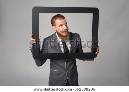 Hipster business man with beard and mustashes in suit looking through peeping out of frame, over grey background