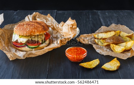 Fast food restaurant dish top view. Meat cheese burger in craft paper, potato chips and wedges. Take away set on dark black wood background. Hamburger and spicy tomato sauce.