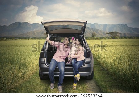Happy family sitting behind the car while taking selfie picture using smartphone in the rice field 