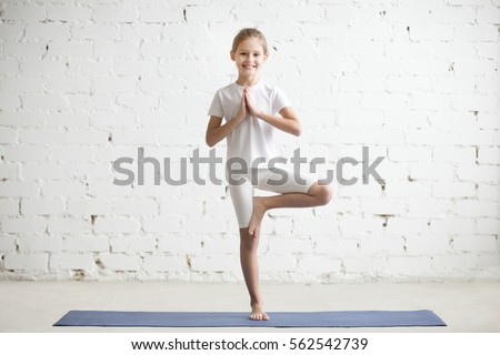 Smiling girl child practicing yoga, standing in Vrksasana exercise with namaste, Tree pose, working out wearing sportswear, t-shirt, pants, indoor full length, white loft studio background  Royalty-Free Stock Photo #562542739