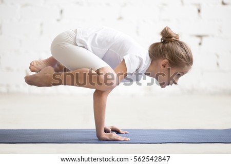 Side view of Girl child practicing yoga, standing in Crane exercise, Bakasana pose, working out wearing sportswear, t-shirt, pants, indoor full length, white loft studio background  Royalty-Free Stock Photo #562542847