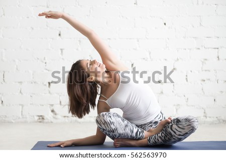 Young cheerful attractive woman practicing yoga, sitting in Sukhasana pose, side bend exercise, working out wearing sportswear, top, pants, indoor full length, white loft studio background  Royalty-Free Stock Photo #562563970