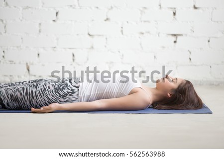 Young attractive woman practicing yoga, lying in Dead Body, Savasana exercise, resting in Corpse pose, working out wearing sportswear, indoor, white loft studio background, closeup  Royalty-Free Stock Photo #562563988