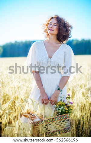 smiling beautiful woman in white summer dress in a field among the wheat ears and holding a basket with bread, milk and a bouquet of wildflowers