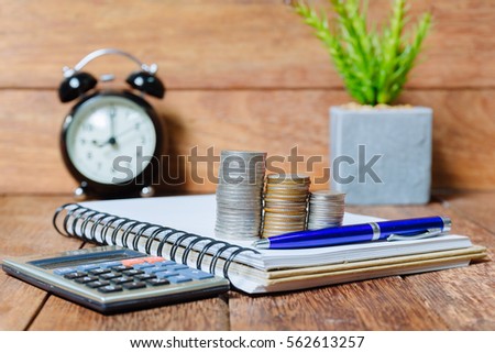 pile of money coins with alarm clock and pen and calculator and notebook on wood background