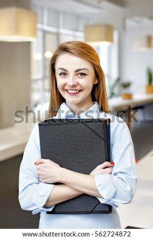 Portrait of young happy smiling businesswoman with black folder in hands in a light office
