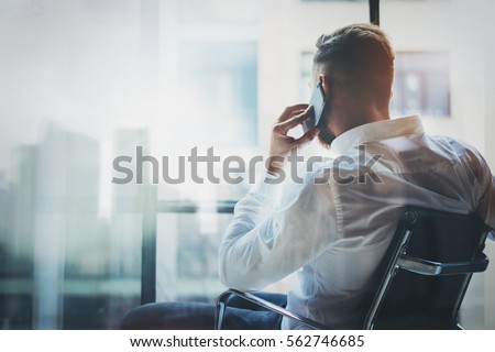 Stylish banker making mobile call at modern loft after work day.Man sitting in chair and looking out the panoramic window.Skyscraper office building blurred on background.Horizontal Royalty-Free Stock Photo #562746685