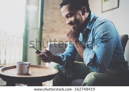 Smiling bearded African man using smartphone while sitting on sofa at home.Concept people working with mobile gadget.Blurred background Royalty-Free Stock Photo #562748419