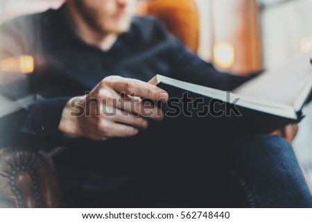 Closeup view of male hands holding book.Young bearded man relaxing at home while sitting in vintage chair.Selective focus on hand,blurred background.Horizontal, film effect