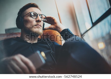 Portrait handsome bearded man wearing glasses,headphones listening to music at modern home.Guy sitting in vintage chair,holding smartphone and relaxing.Panoramic windows background.Blurred background Royalty-Free Stock Photo #562748524