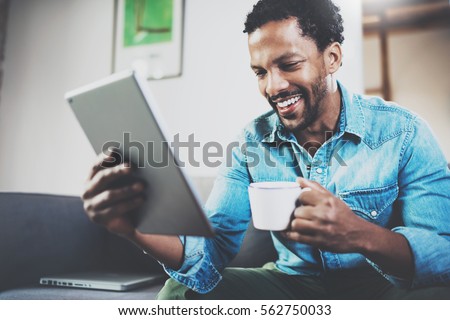 Young bearded African man using tablet while sitting on sofa and holding white cup coffee in hand at home.Concept people working with mobile gadget.Blurred background Royalty-Free Stock Photo #562750033