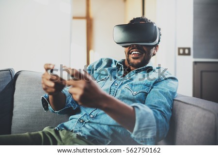 Bearded african man enjoying virtual reality glasses while sitting on sofa.Happy young guy with vr headset or 3d spectacles and controller gamepad playing video game at home.Blurred
