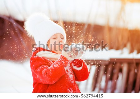 little cute girl 4 -5 years in a red coat, white hat with a fluffy pompon walks in the winter in the street blowing a palm full of snow, the snow is flying beautifully in the rays of the setting sun