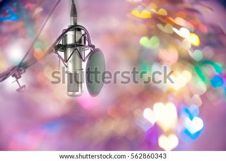Condenser microphone with pop filter windscreen on stand ready for recording on stage in party event .
High fidelity microphone on stage with colourful heart  bokeh background .