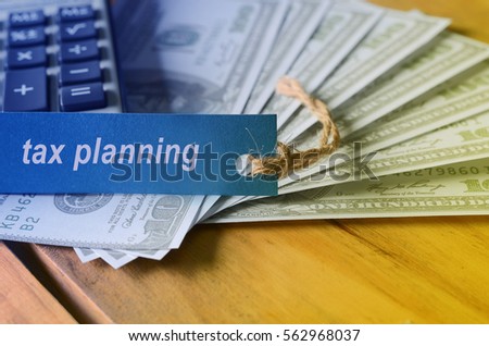 Finance conceptual image with TAX PLANNING words, hundred dollar bills, coins in a jar, a magnifying glass and calculator on wooden background. tone image.