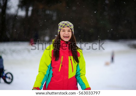 Winter portrait of the young woman in a bright ski suit and a knitted cap. It is snowing. Cute woman rejoices to winter and snow as the child.