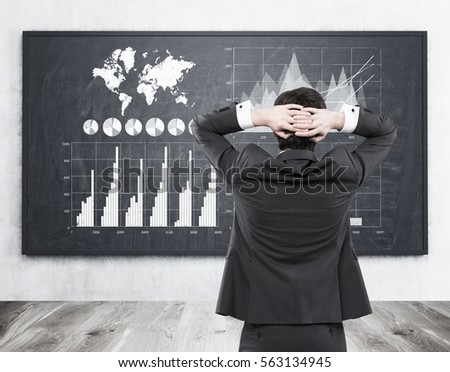Rear view of a businessman standing with his arms behind his head and looking at white and blue graphs on a blackboard. Elements of this image furnished by NASA