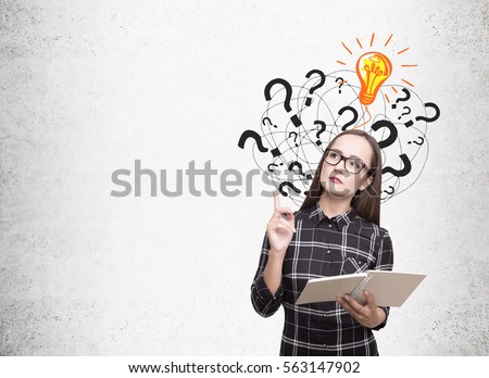 Nerdy girl in glasses and a checkered shirt is holding a book and standing near a concrete wall with light bulb and question marks. Mock up