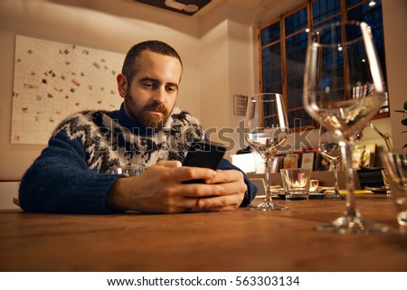 Thoughtful bearded man in warm wool knitted icelandic sweater with horses using his smart phone, sitting in served table for holiday dinner, warm lit ambient lightning inside big spacious room