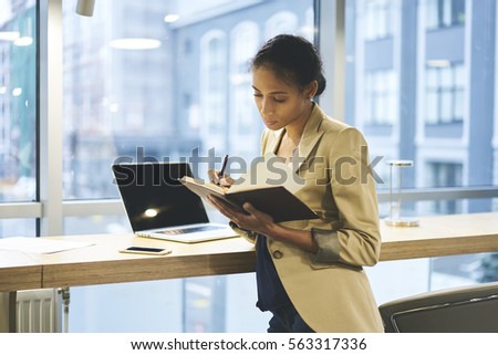 Attractive female manager doing remote paperwork making notes in personal diary using technology and wireless connection to internet standing near copy space area for your advertising content or text