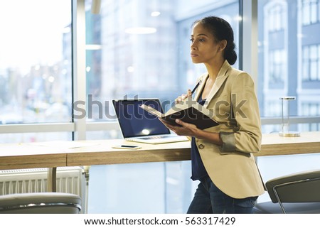 Serious formal dressed attractive afro american female administrative making notes to organizer using laptop computer with mock up screen and internet in office copy space for your advertising
