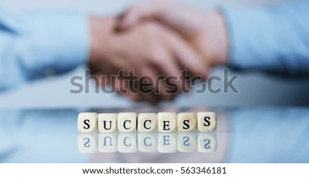 Slow motion macro of the word "success" made of the wooden letter blocks and behind a handshake of two persons in shirt conclude the deal. Concept: affair, business, finance, money.
