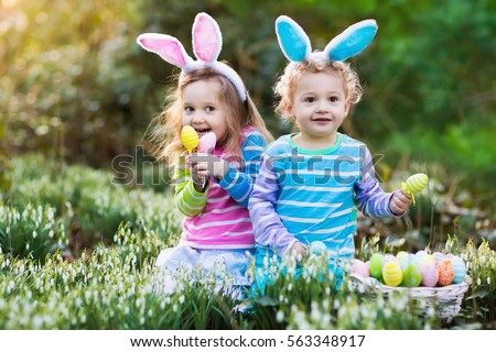 Kids on Easter egg hunt in blooming spring garden. Children with bunny ears searching for colorful eggs in snow drop flower meadow. Toddler boy and preschooler girl in rabbit costume play outdoors
