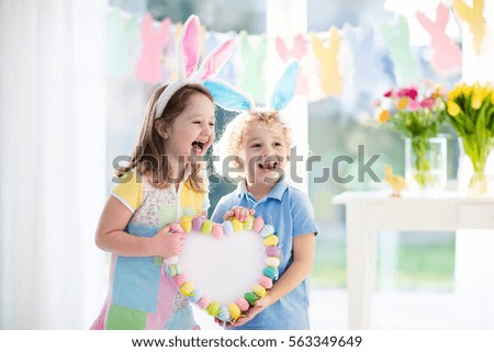 Little boy and girl in bunny ears holding a heart frame with colorful Easter eggs. Kids celebrate Easter. Children having fun on Easter egg hunt. Pastel bunny banner and flowers. Space for your text