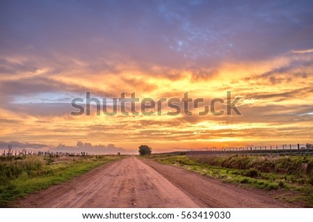 Dust road in countryside of Argentina, orange colorful clouds, panoramic scene. Open horizon view, grass, farm fence at the side