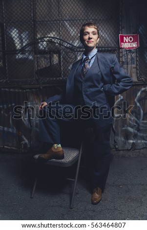 Young androgynous woman dressed in mens clothing in a grungy, urban outdoor location stands confidently