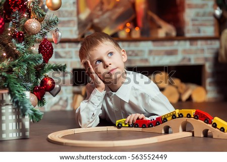 Cute blond boy of 6 years in a white shirt plays with wooden railway, in New Year room near the fireplace and the beautiful large Christmas tree decked out with gold, red and silver toys