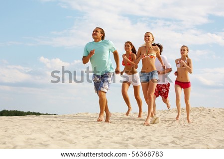 Photo of happy friends running down sandy beach on background of cloudy sky