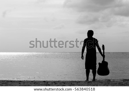 Silhouette of young man holding his guitar while standing up looking out to the sea at sunset in the island of Koh Phangan, Thailand. Music artist finding inspiration. Black and white photography