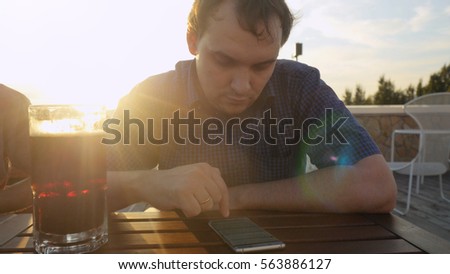 Young man with smartphone eating and drinking beer in outdoor cafe during sunset  lense flare effects
