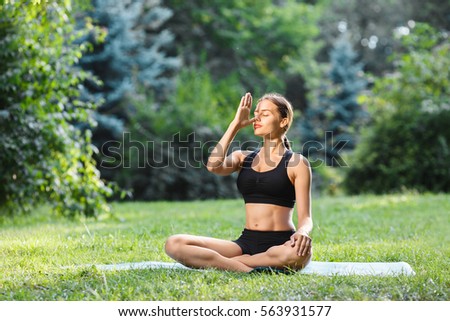 Young woman with brown hair wearing black top and shorts doing yoga position in park, healthy lifestyle, portrait, pranayama. Royalty-Free Stock Photo #563931577