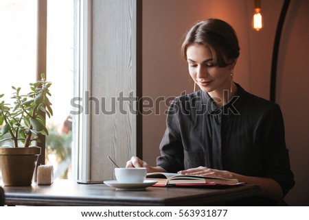 Photo of gorgeous young lady sitting at the table reading book in cafe and looking aside while drinking coffee.