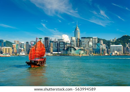 Traditional Chinese wooden sailing ship with red sails in Victoria harbor in rays of setting sun. Skyscrapers in downtown of Hong Kong are visible from Kowloon side. The Hong Kong Island skyline.