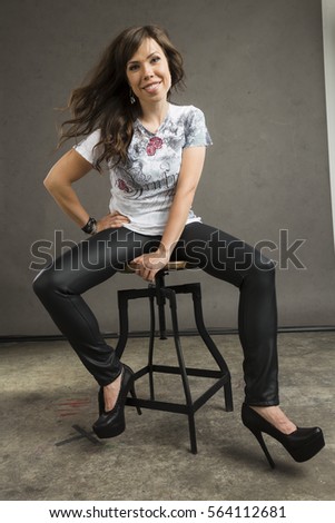 Young caucasian woman with brown hair, a grey top, leather pants, and black high heels sits on a metal stool	