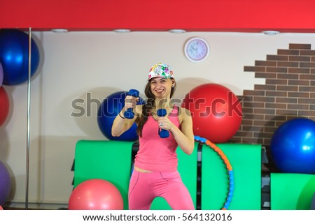 Young beautiful white girl in a pink sports suit with dumbbells in her hands before training in a fitness club. Interior of fitness club for background.