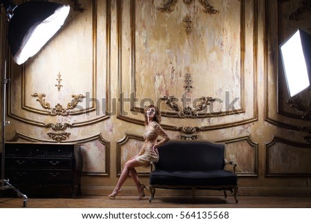 Actress in a short shiny dress with sequins sitting on the sofa armrest in front of studio light in an ancient palace interior