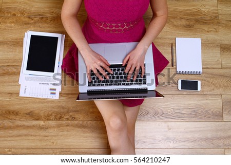 Girl in purple dress works on laptop, sitting on the wooden floor with the tablet, smartphone, notepad and papers near, top view, freelancer concept