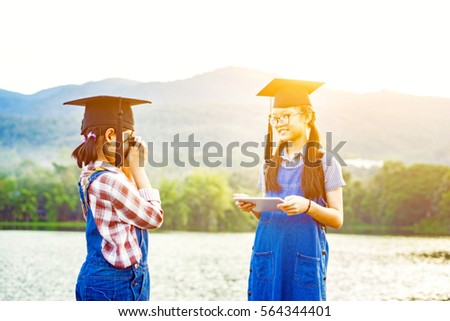 Two young girls graduate being photograph in a park with a DSLR mirrorless camera,happy international students in boards and bachelor gowns taking photo with smartphone ,morning light,vintage color.