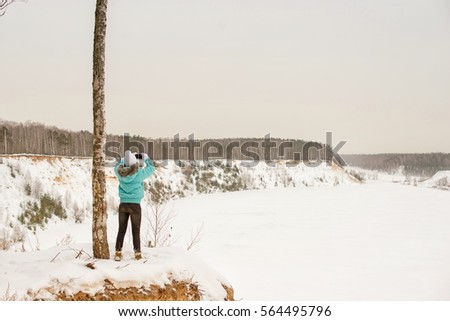 Girl in bright clothes on the edge of the cliff on the background of the winter forest in winter. In the hands of the phone, take pictures on the phone.