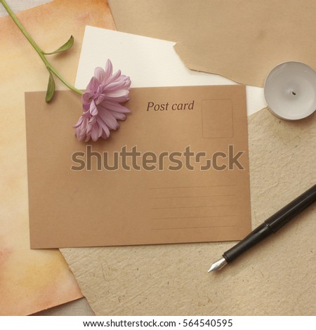 A square photo of a vintage style still life with a brown paper postcard with a place for text, a tender pink chrysanthemum, an ink pen, a candle, and some old papers, shot from above