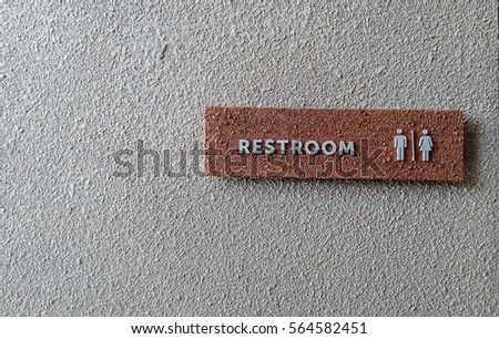 Restroom sign is made from red stone which has grey alphabets on in. It is attached with grey wall. This photo was taken from wide angle.