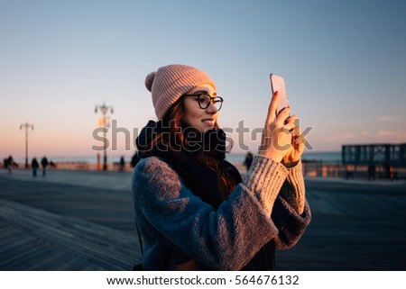 Portrait of a smiling young woman in eyeglasses taking picture with mobile phone at the sea pier